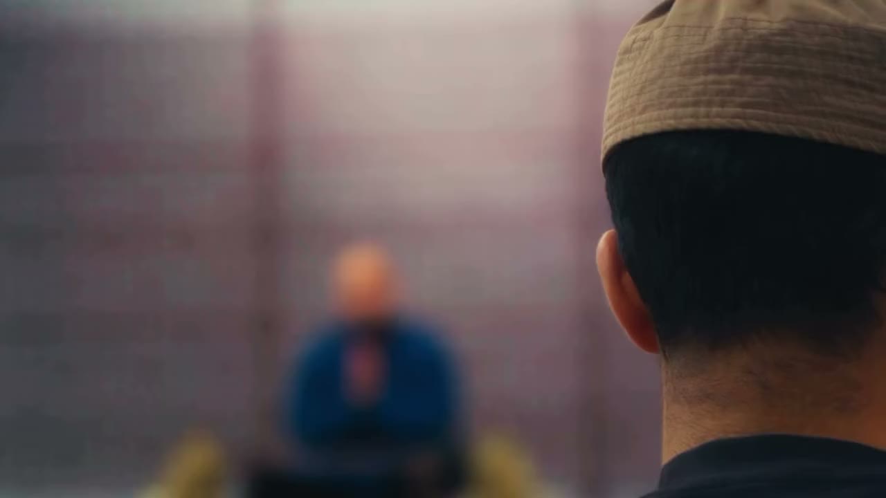 Focus shifts from a man in a kufi to Sh. Ahmed Billoo seated before a lattice screen, then a wide shot of him speaking with open hands.