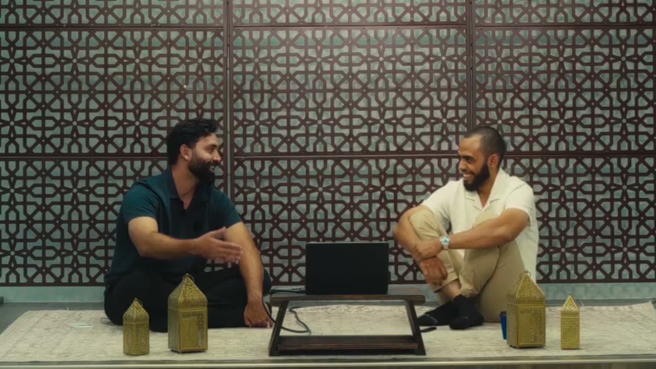 Two men on a rug in front of a lattice wall: one leans back laughing while the other talks with open hands.