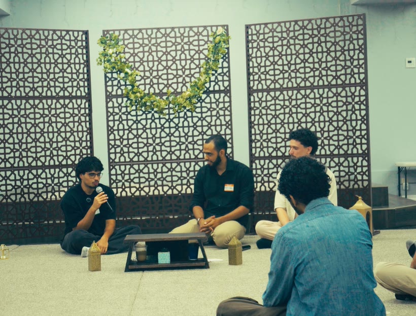 A fireside circle on floor seating in front of lattice screens and a garland, with an audience member seen from behind in the foreground.