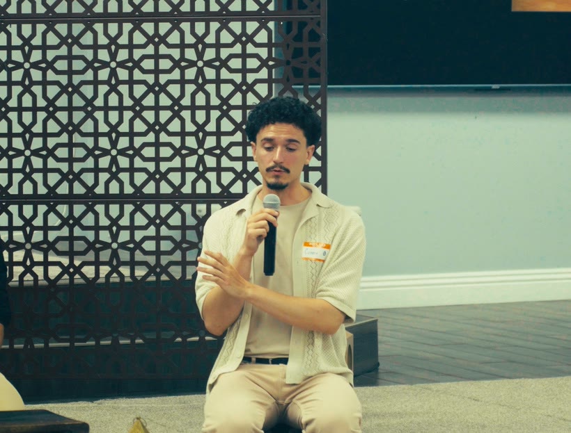 A speaker with a handheld microphone gestures while seated, in front of a lattice screen.