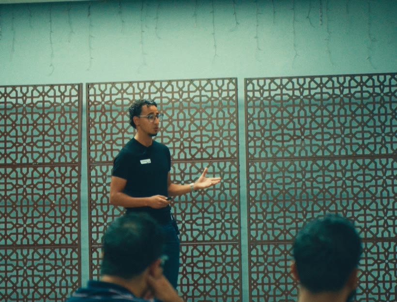 A speaker stands in front of a lattice wall, gesturing with one hand, with audience heads in the foreground.