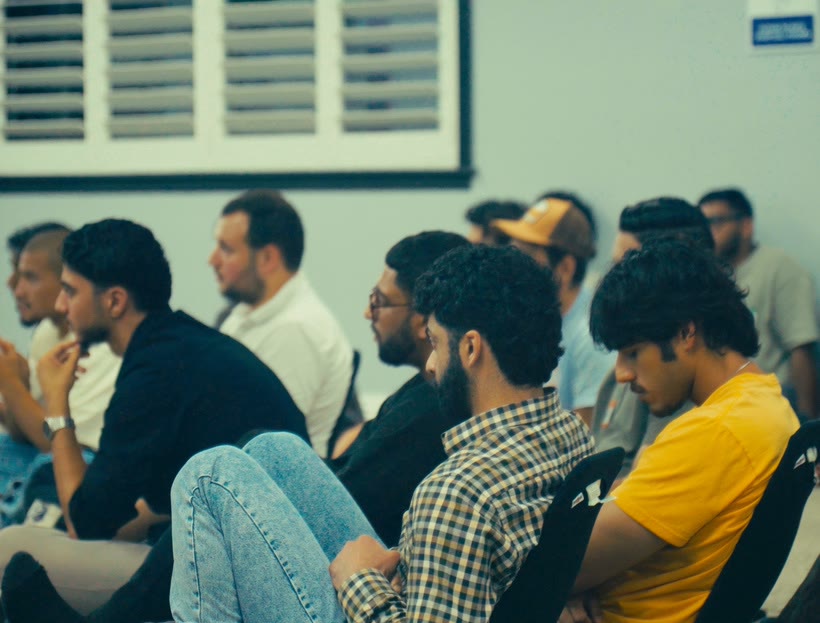 Close-up of seated attendees in profile, listening, in a bright room with shuttered windows.