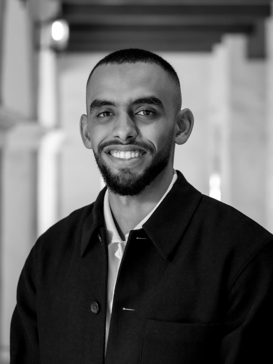 Portrait of Ahmed Hageb, smiling, in a black overcoat over a white collar, standing in a sunlit arcade with wooden ceiling beams.