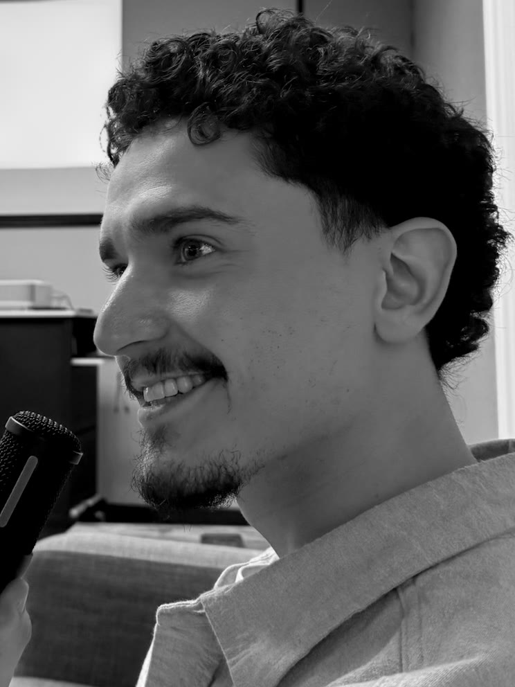 Portrait of Connor Gonzales smiling in profile, holding a microphone, seated on a couch.