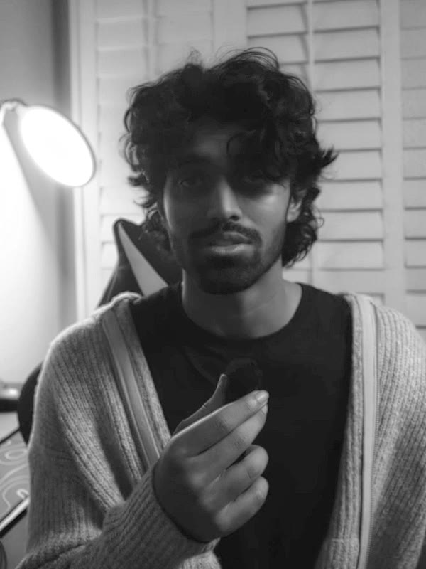 Portrait of Eshan Parveez in a black T-shirt and a light knit cardigan, mid-gesture, with white shutters and a lamp behind him.
