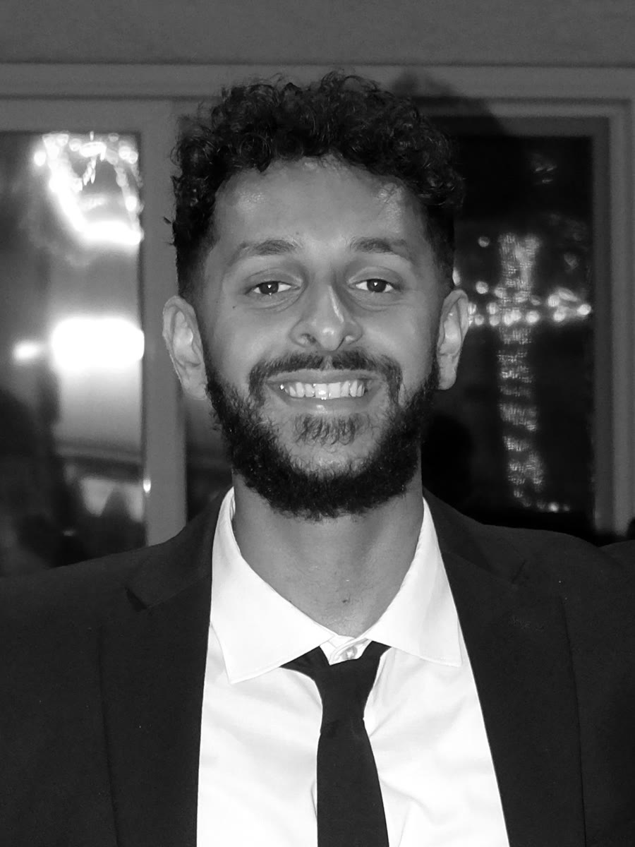 Portrait of Usman Qazi smiling at the camera in a black suit and tie, with string lights reflected in the window behind him.